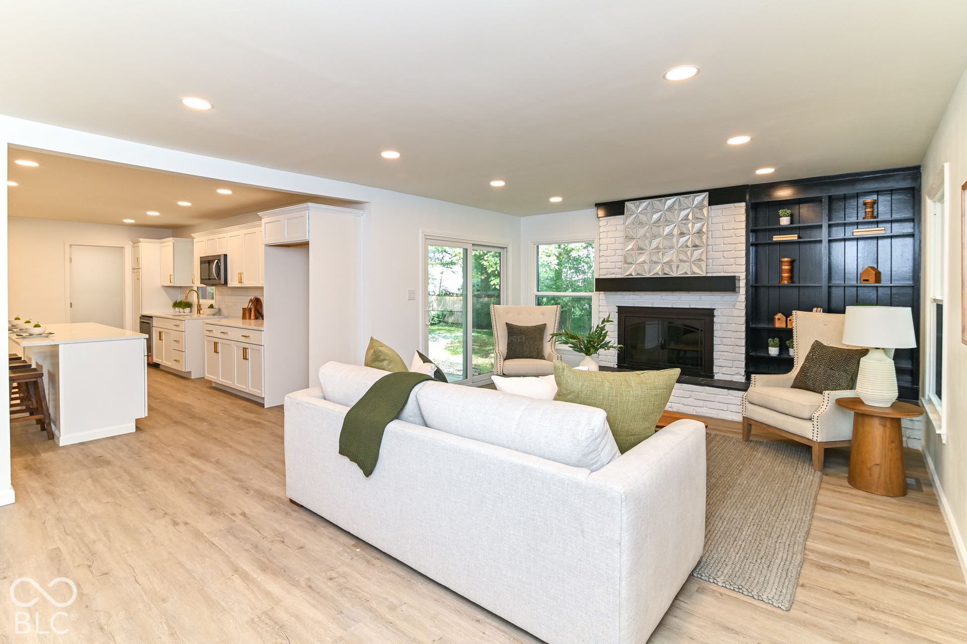 Open-concept living area featuring wood-finish flooring, a white brick fireplace with a dark mantel, built-in shelving, and recessed lighting
