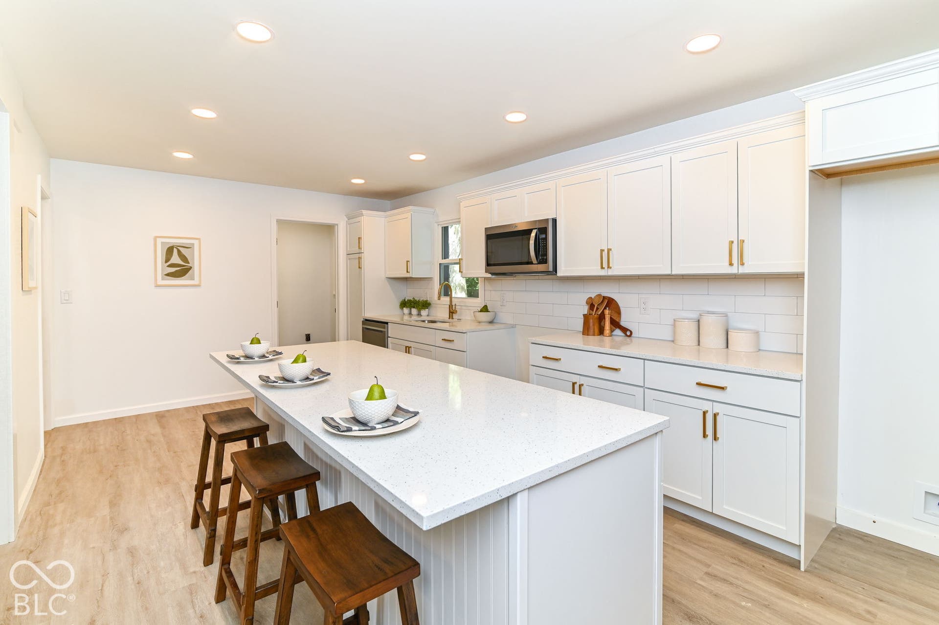 Spacious kitchen featuring an extended island with a light-toned countertop, white cabinetry with gold-toned hardware, stainless steel microwave, white subway tile backsplash, and wood-finish flooring