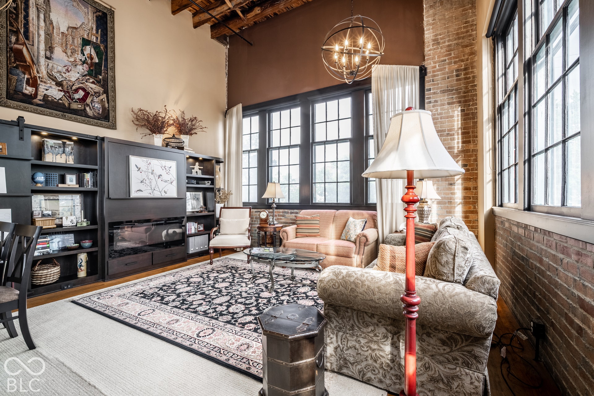 Exposed brick wall, high ceilings with exposed wood beams, tall multi-pane windows, built-in entertainment center with shelving, and overhead spherical chandelier