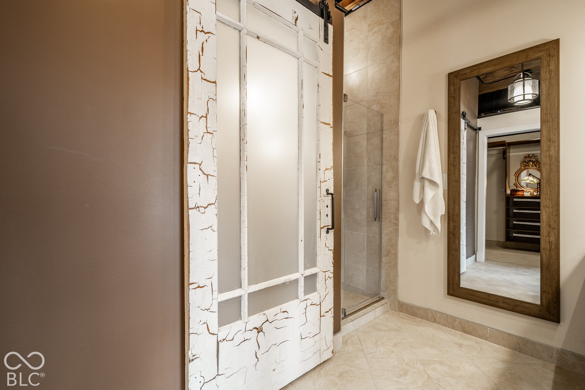 Frosted glass paneled barn door, neutral-toned wall paint, full-length framed mirror, glass-enclosed shower, and light-toned tile flooring