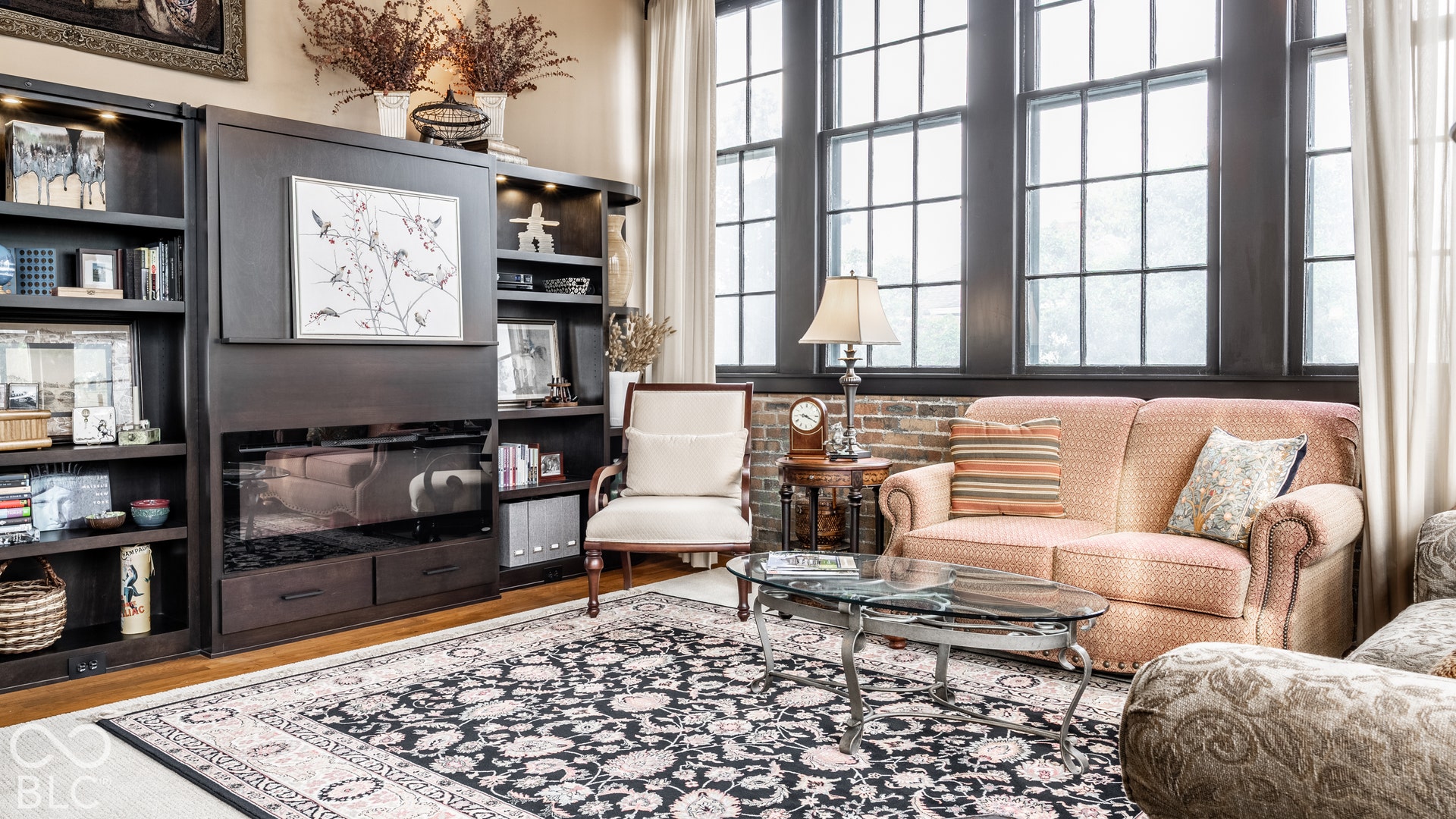 Built-in shelving unit with integrated lighting, exposed brick wall feature, large multi-pane windows, and wood-finish flooring