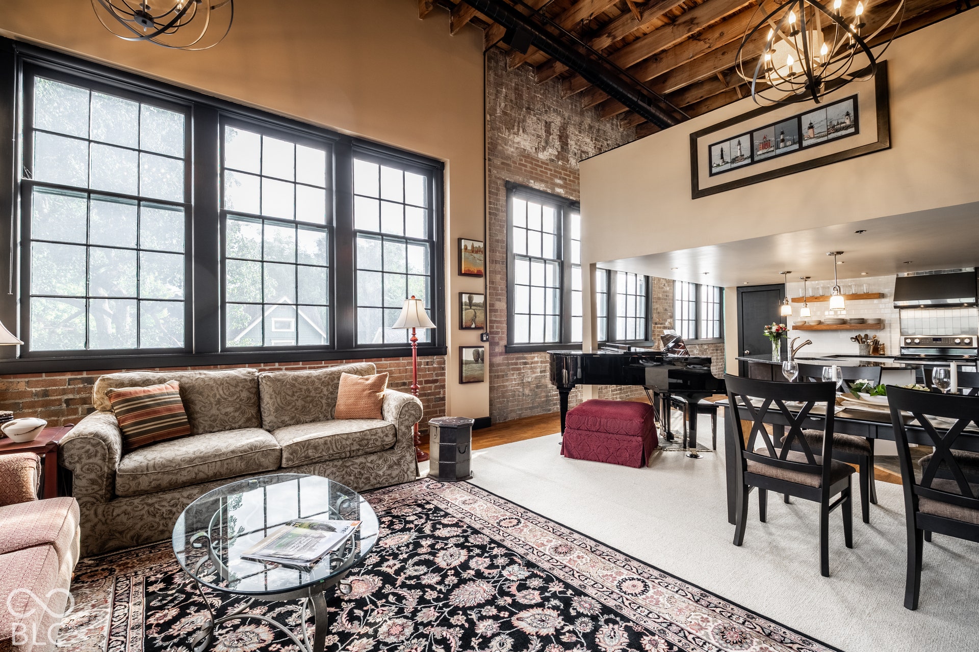 Exposed brick wall, high ceilings with exposed wood beams, large grid-pattern windows, open-concept layout, and a contemporary kitchen with a tiled backsplash