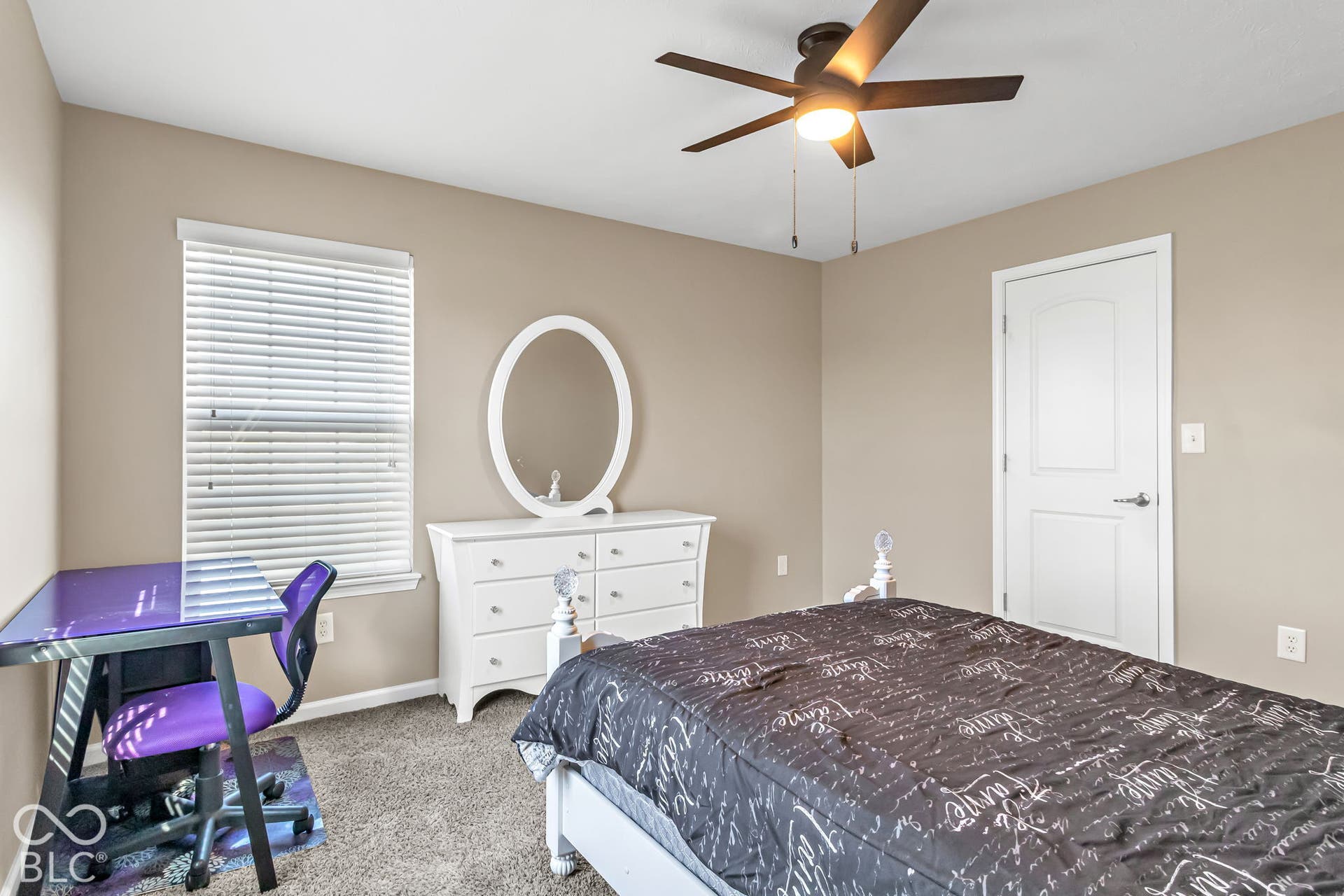 Textured carpet, neutral wall color, window with horizontal blinds, ceiling fan with integrated lighting, and a white paneled door
