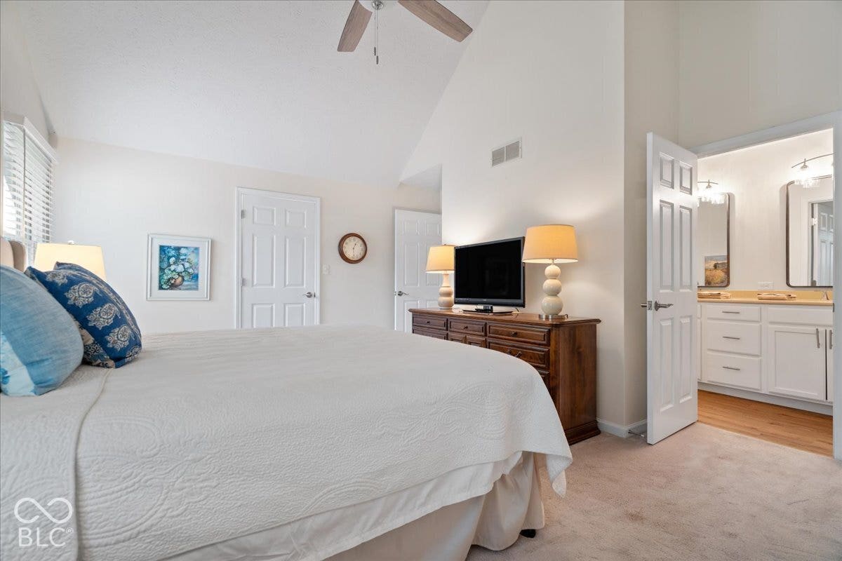 Vaulted ceiling with a centrally mounted ceiling fan, neutral wall-to-wall carpeting, and an attached bathroom featuring white cabinetry with integrated countertop sink and framed wall mirrors.