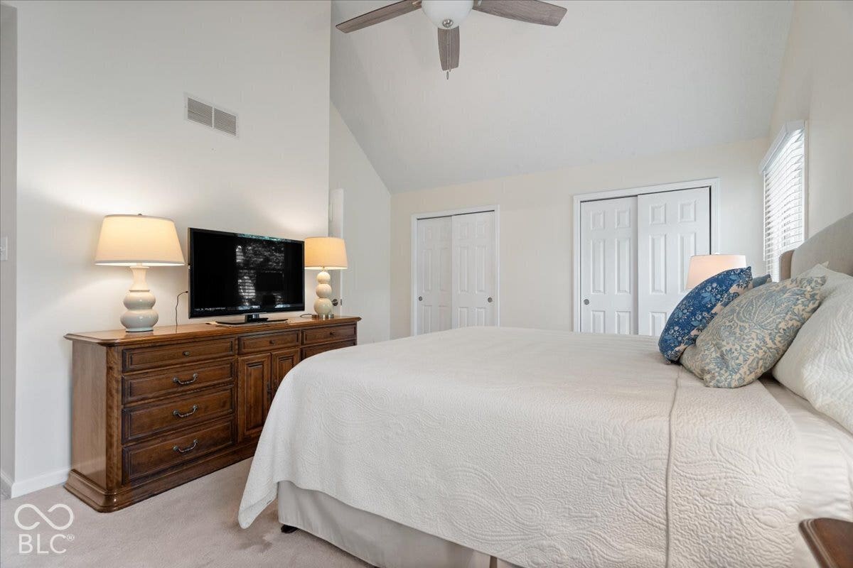 Vaulted ceilings, neutral carpeting, dual closets with raised-panel doors, and a ceiling-mounted light fixture with fan. Double closets with access to attic storage.
