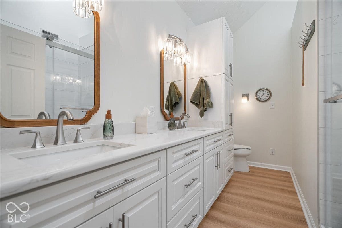Updated Double vanity with white cabinetry, stone-finish countertops, dual rectangular undermount sinks, wood-finish flooring, and arched wood-frame mirrors