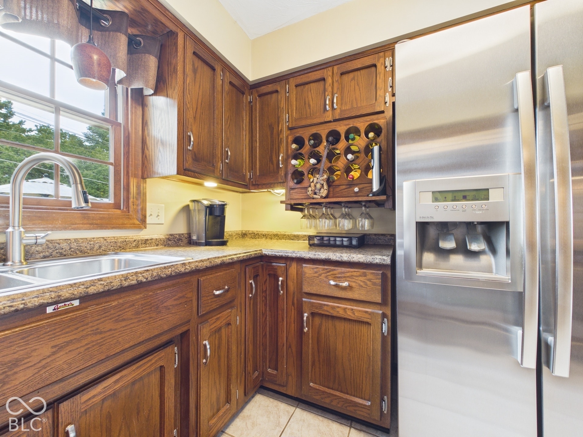 Wood-finish cabinetry with silver-tone hardware, stainless steel refrigerator, double basin sink with gooseneck faucet, built-in wine rack, and under-cabinet lighting