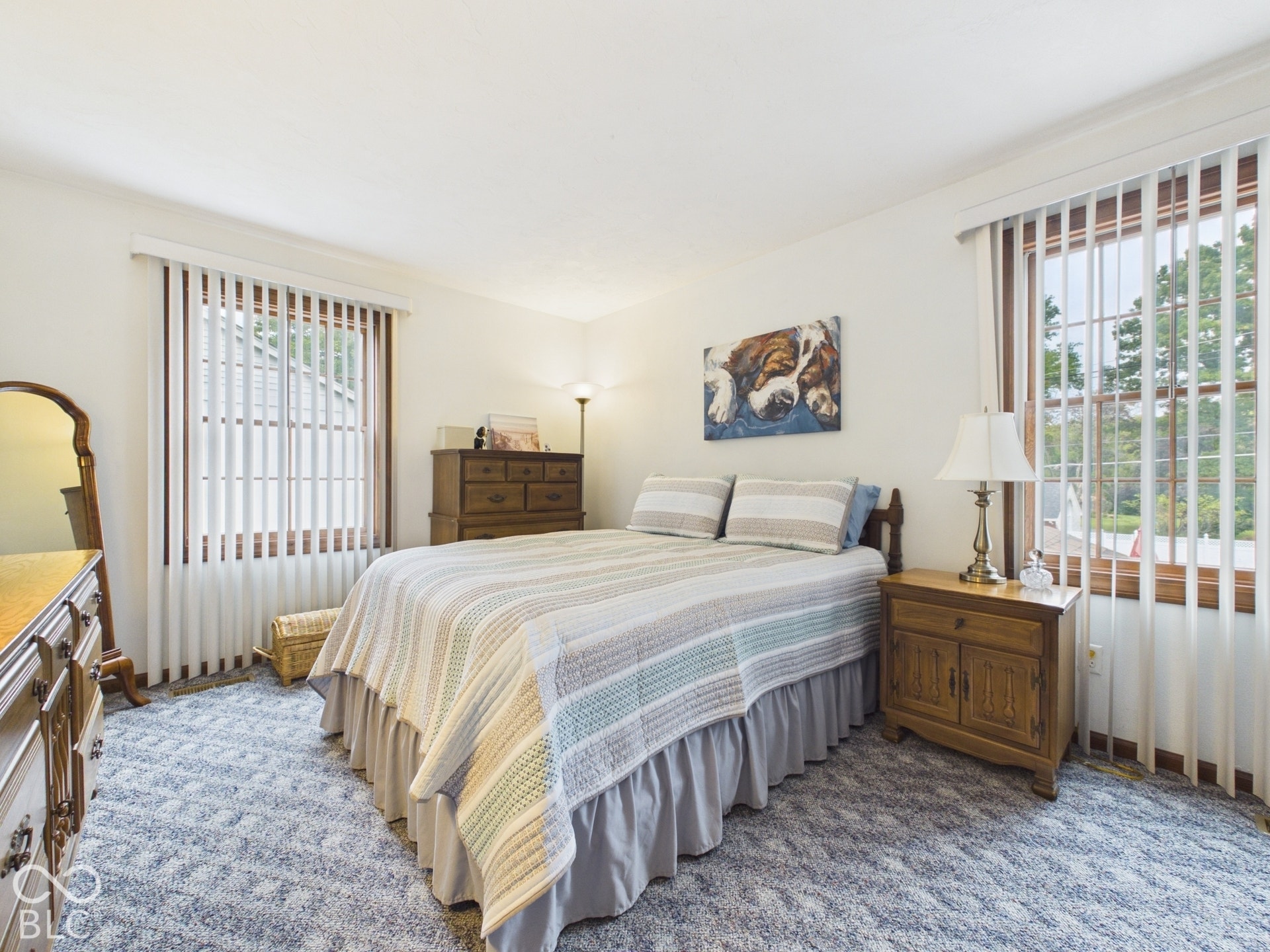 Light-colored wall finishes, two large windows with vertical blinds, and a wood-frame bed