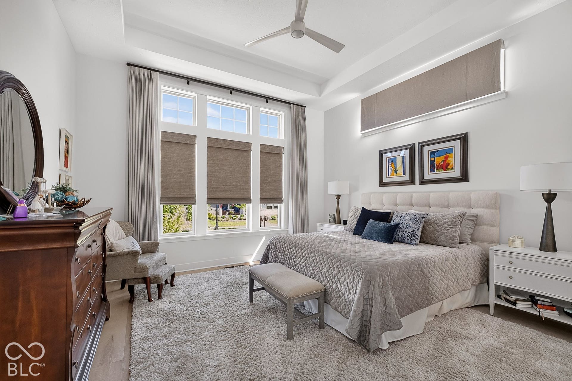 1405 Lake Haven Lane, Westfield, IN 46074 Sophisticated primary bedroom showcasing a coffered ceiling with ceiling fan, expansive windows with cellular shades and layered drapery providing natural light, hardwood flooring throughout, an upholstered headboard, and a wood-finish dresser with attached mirror