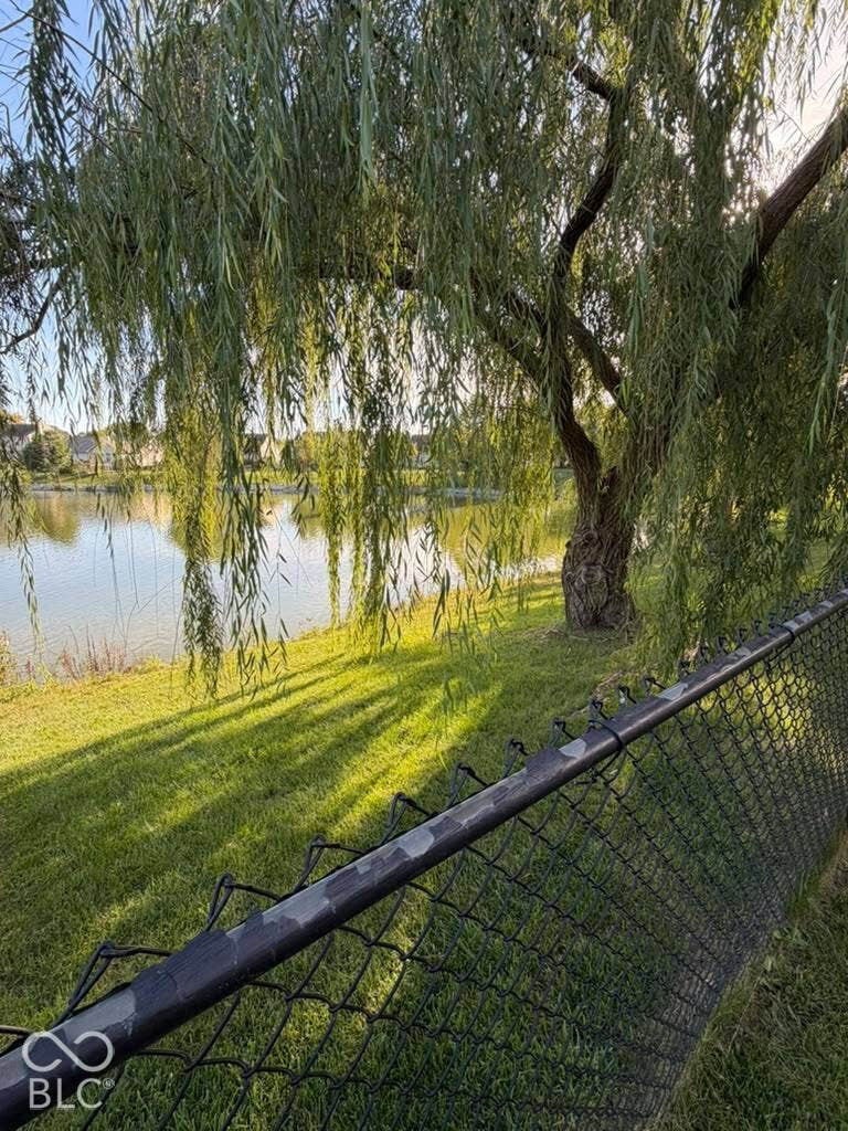 Lakeside property featuring a mature weeping willow tree, manicured lawn, and black chain-link fencing