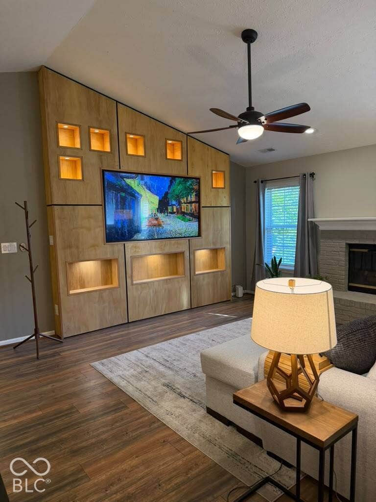 Wood-finish flooring throughout, custom built-in media wall with integrated lighting, ceiling fan with light fixture, and a fireplace with a stone surround