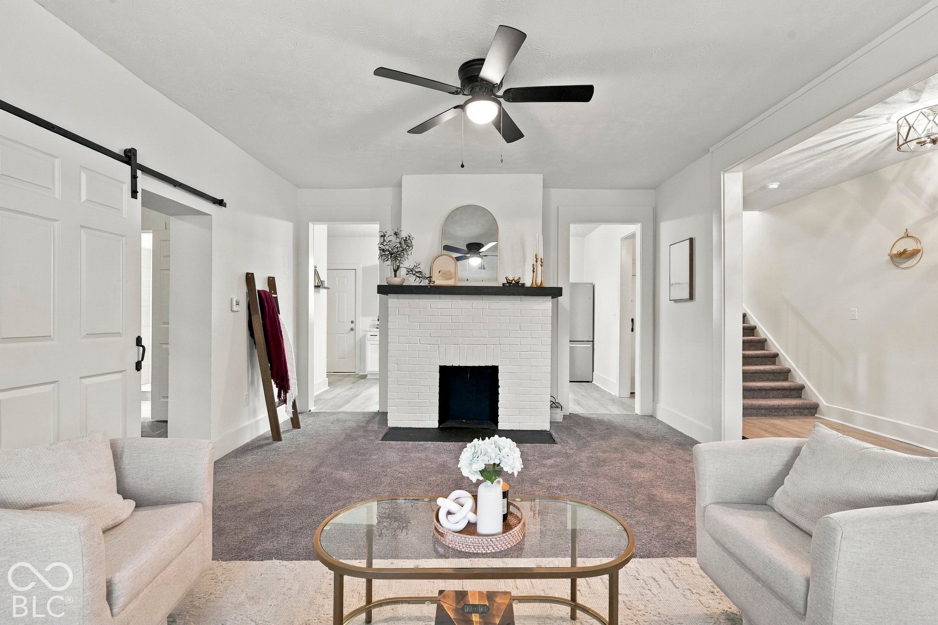 White painted brick fireplace with a dark wood mantel, sliding barn door, ceiling fan with light kit, and carpeted flooring