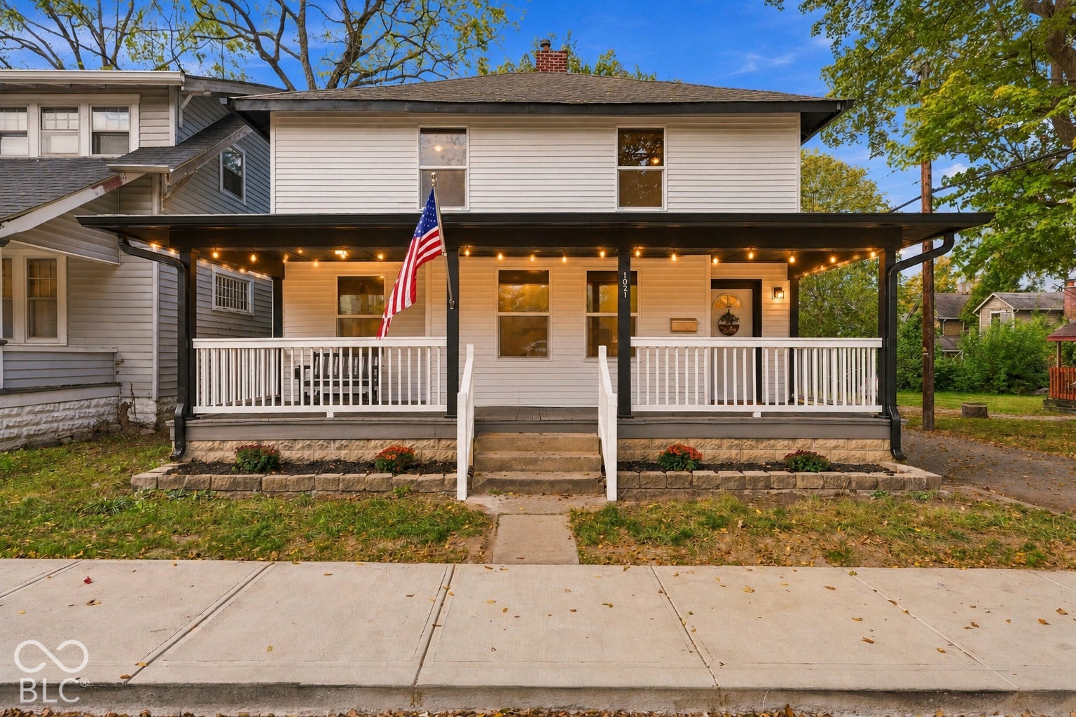 Two-story exterior featuring white horizontal siding, a full-width covered front porch with white railings, a concrete foundation, and a low-profile stone retaining wall