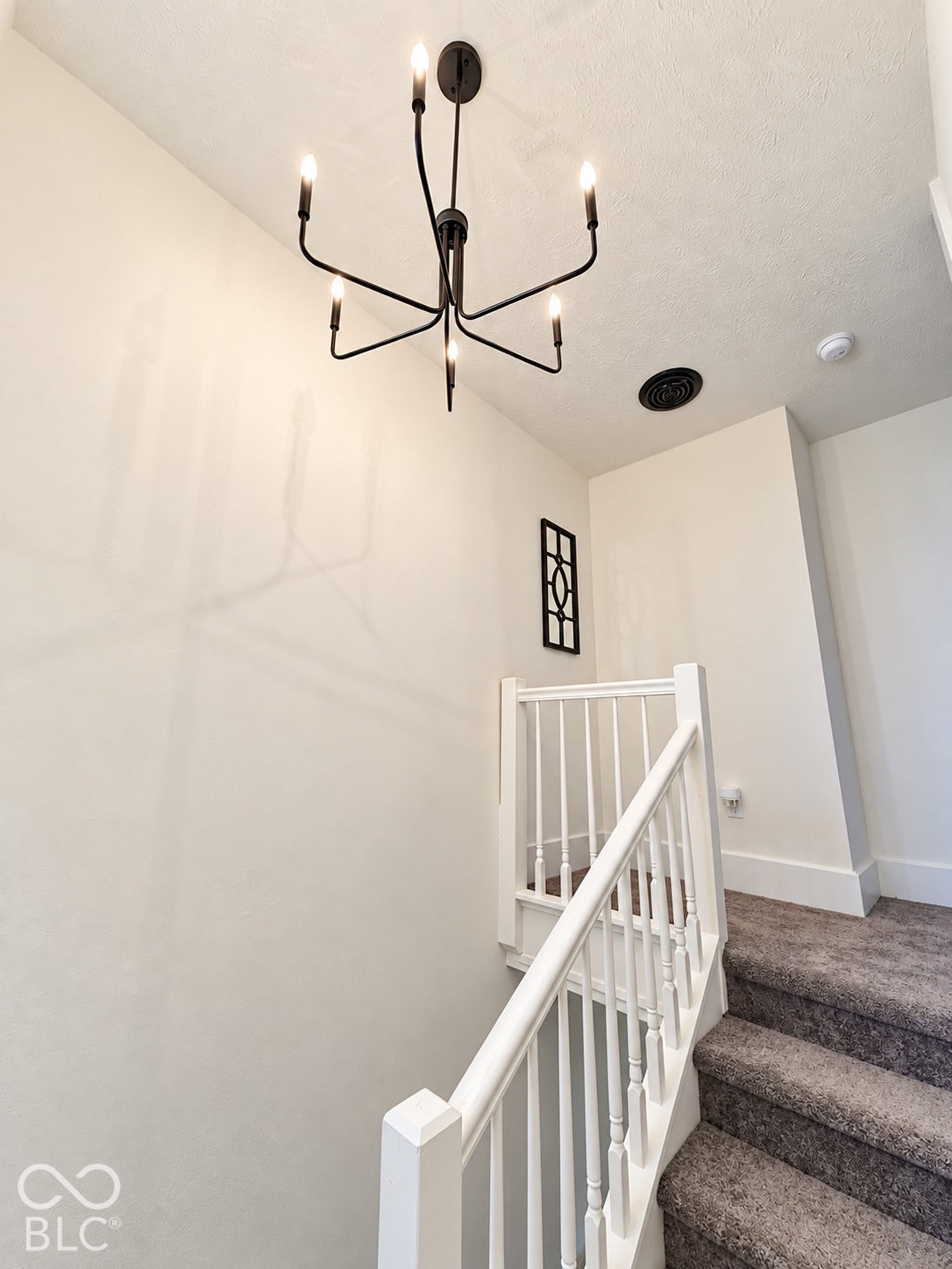 Modern six-arm matte black chandelier, white staircase with baluster railing, and grey carpeting