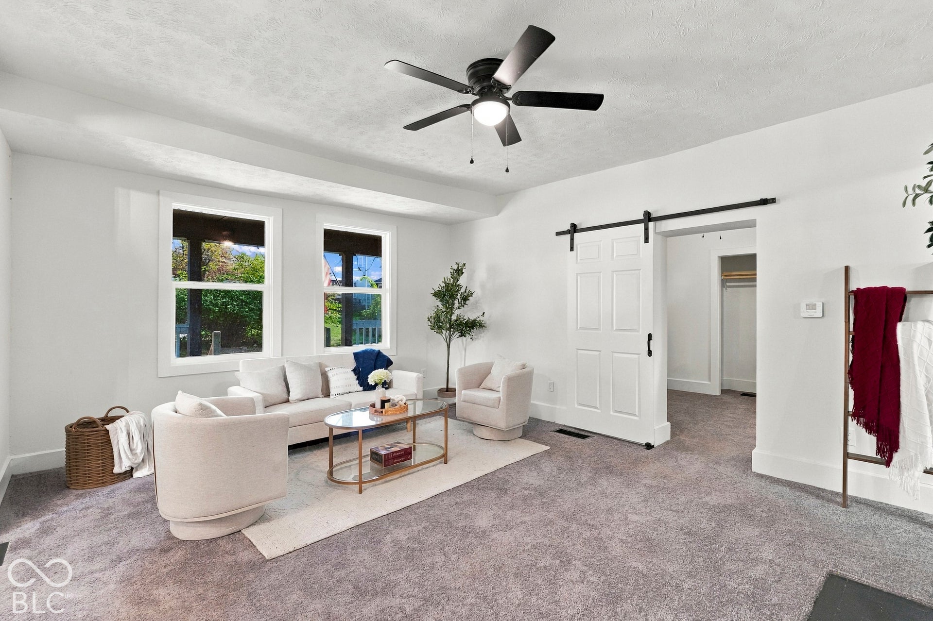 Neutral-toned interior space featuring neutral wall-to-wall carpeting, dual windows with white trim, a black ceiling fan with integrated lighting, and a white paneled sliding barn door