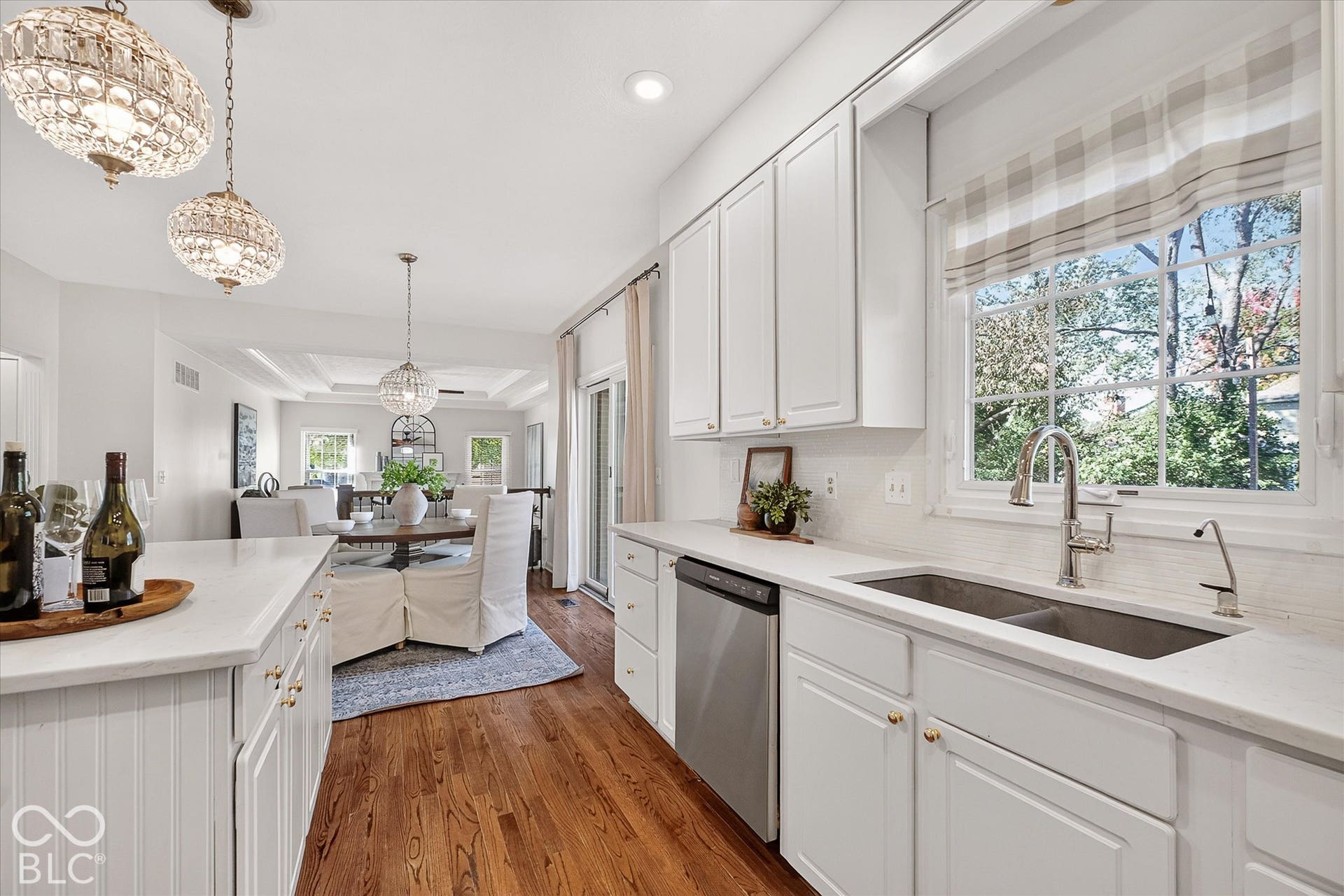 White shaker-style cabinetry with gold-finish hardware, stainless steel double basin sink, white tile backsplash, and wood-finish flooring