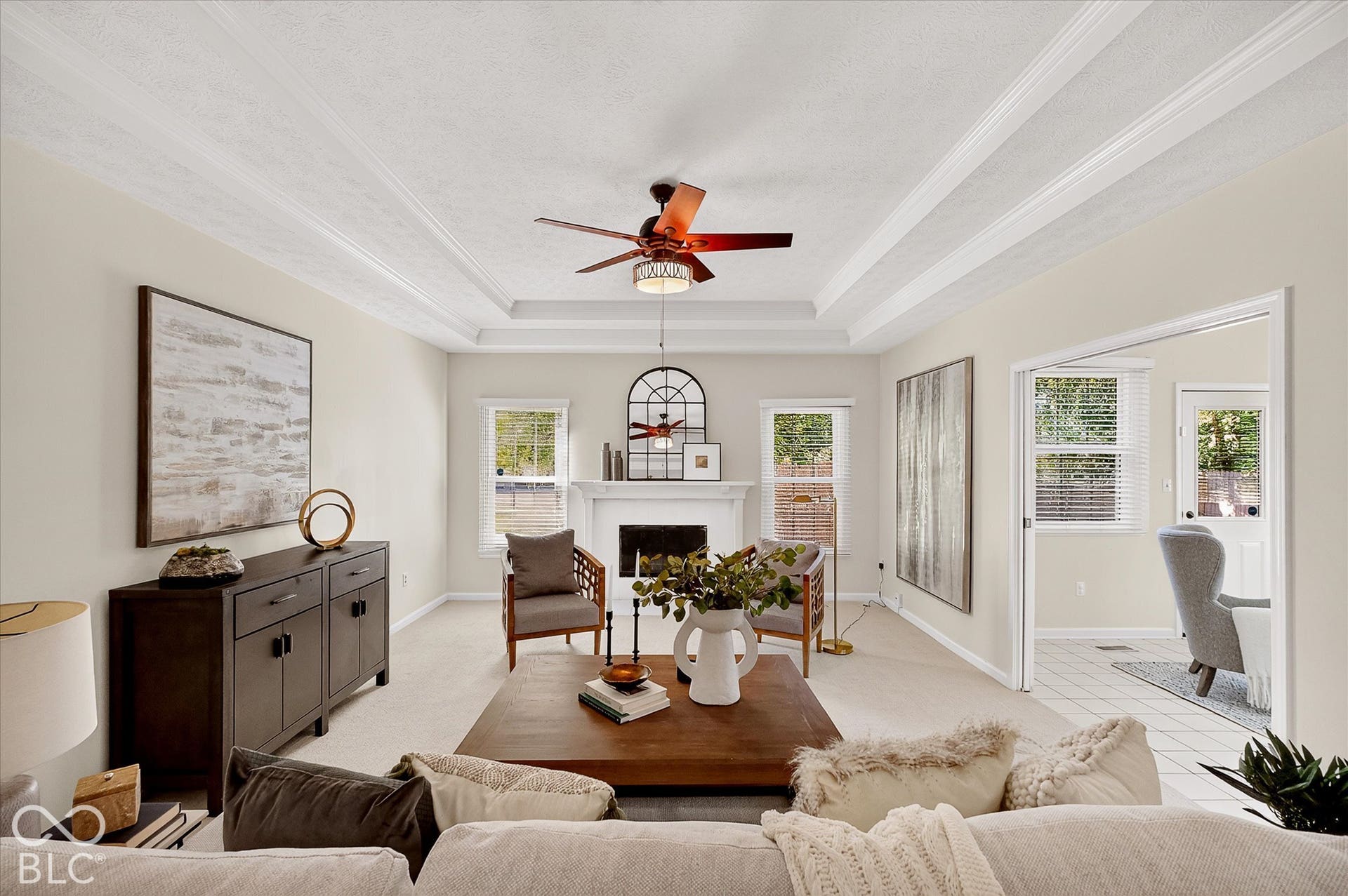 Tray ceiling with decorative molding, ceiling fan, white mantel over fireplace, multiple windows with natural light, and tile flooring visible in the adjacent room