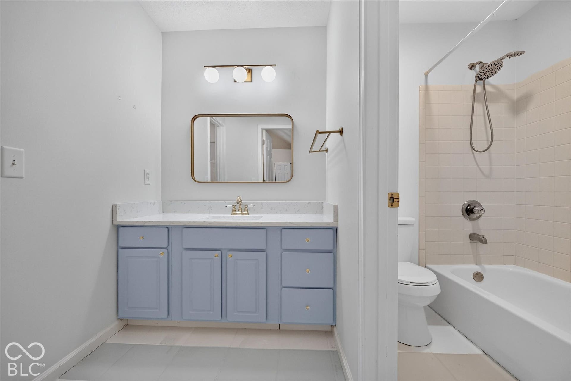 Bathroom featuring a light blue vanity with gold-toned hardware, a rectangular vanity mirror, a three-light vanity fixture, a white countertop, and a white tiled shower and tub combination