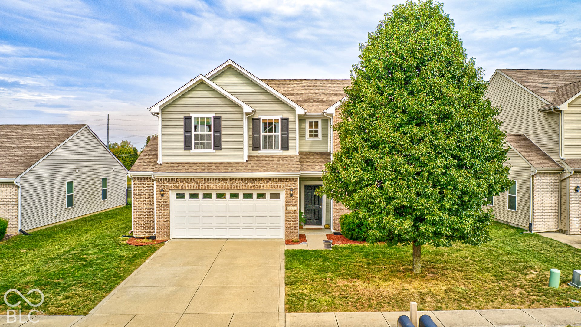 Two-story Traditional American home featuring a brick and siding exterior, two-car garage with paneled doors, and a mature shade tree anchoring the front lawn