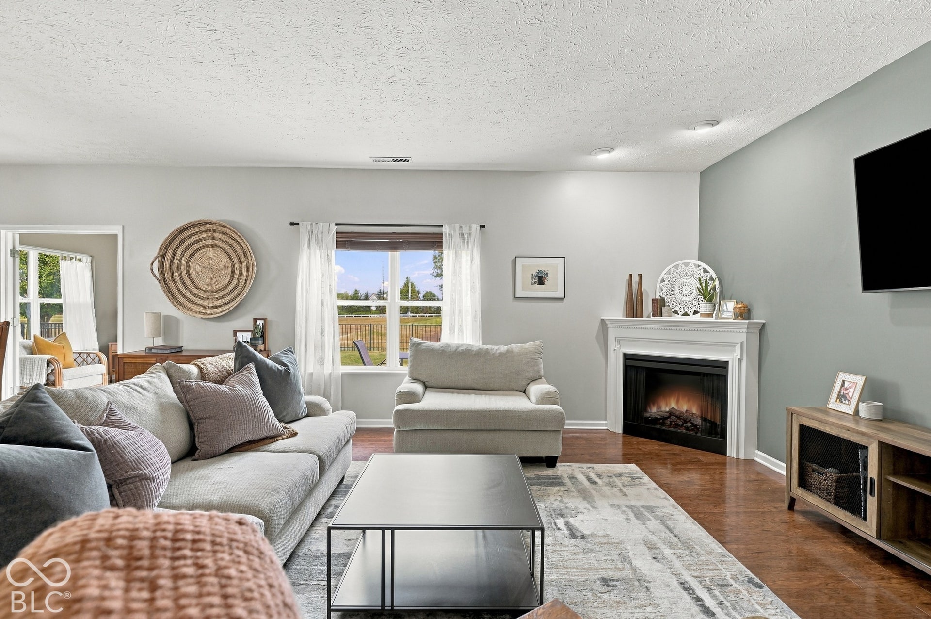 Warm wood-finish flooring, a corner fireplace with a white mantel, and a large window providing abundant natural light