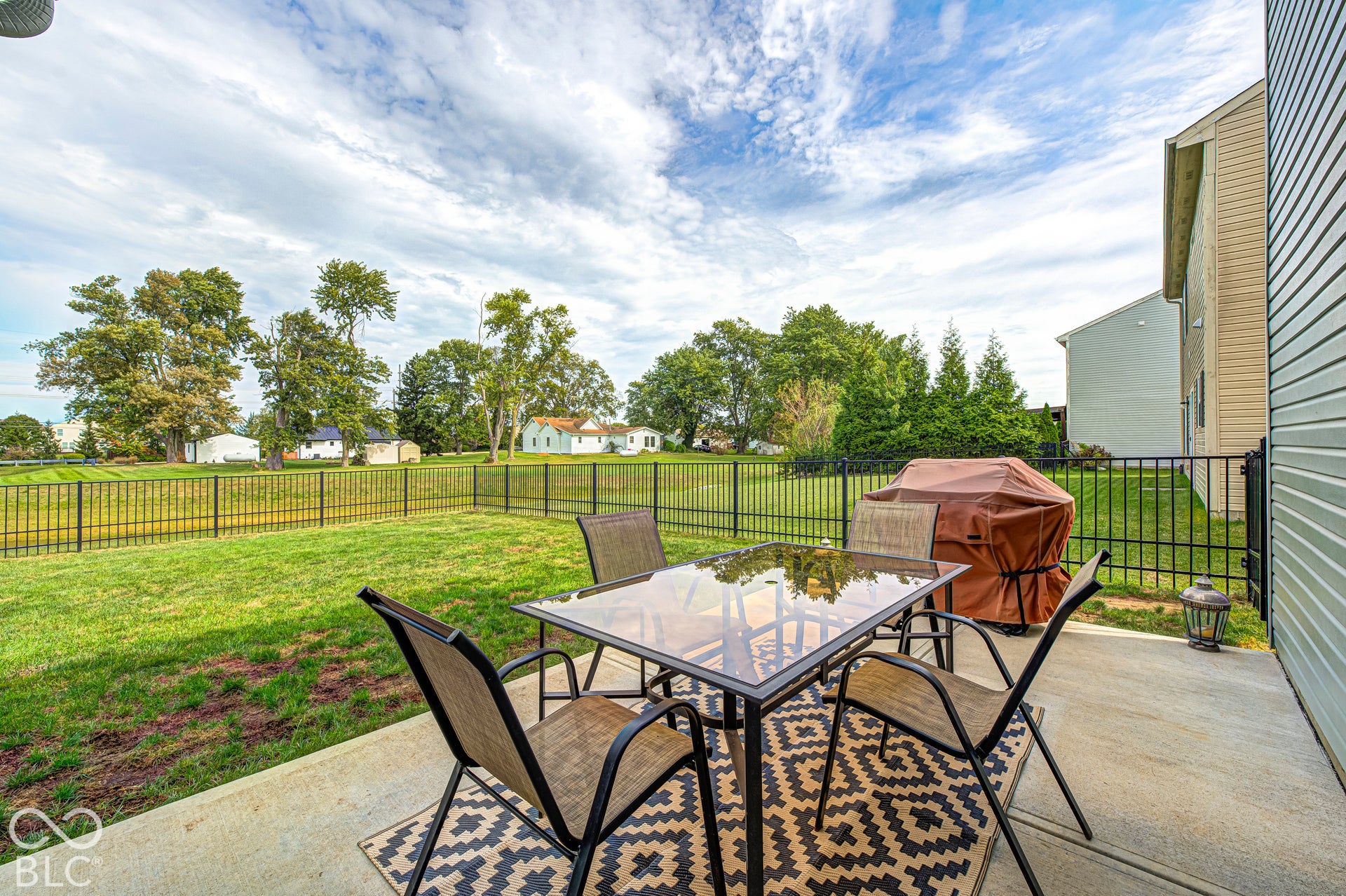 Concrete patio featuring a black metal fence, expansive lawn, and mature trees