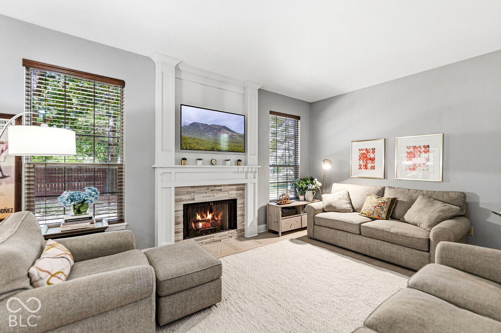 Living area featuring a fireplace with a stacked stone surround and a white mantel, an integrated television alcove, two large windows with blinds, and light-colored carpeting