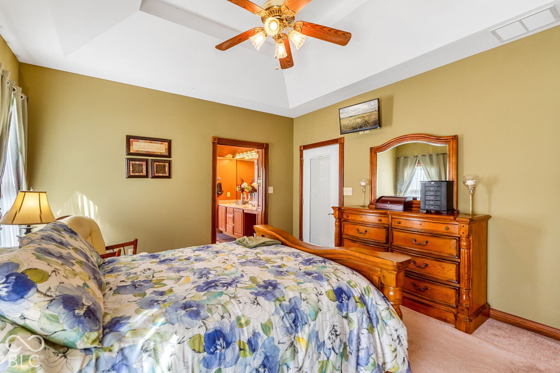 Tray ceiling with a ceiling fan, wall-mounted television, and a doorway leading to an additional space