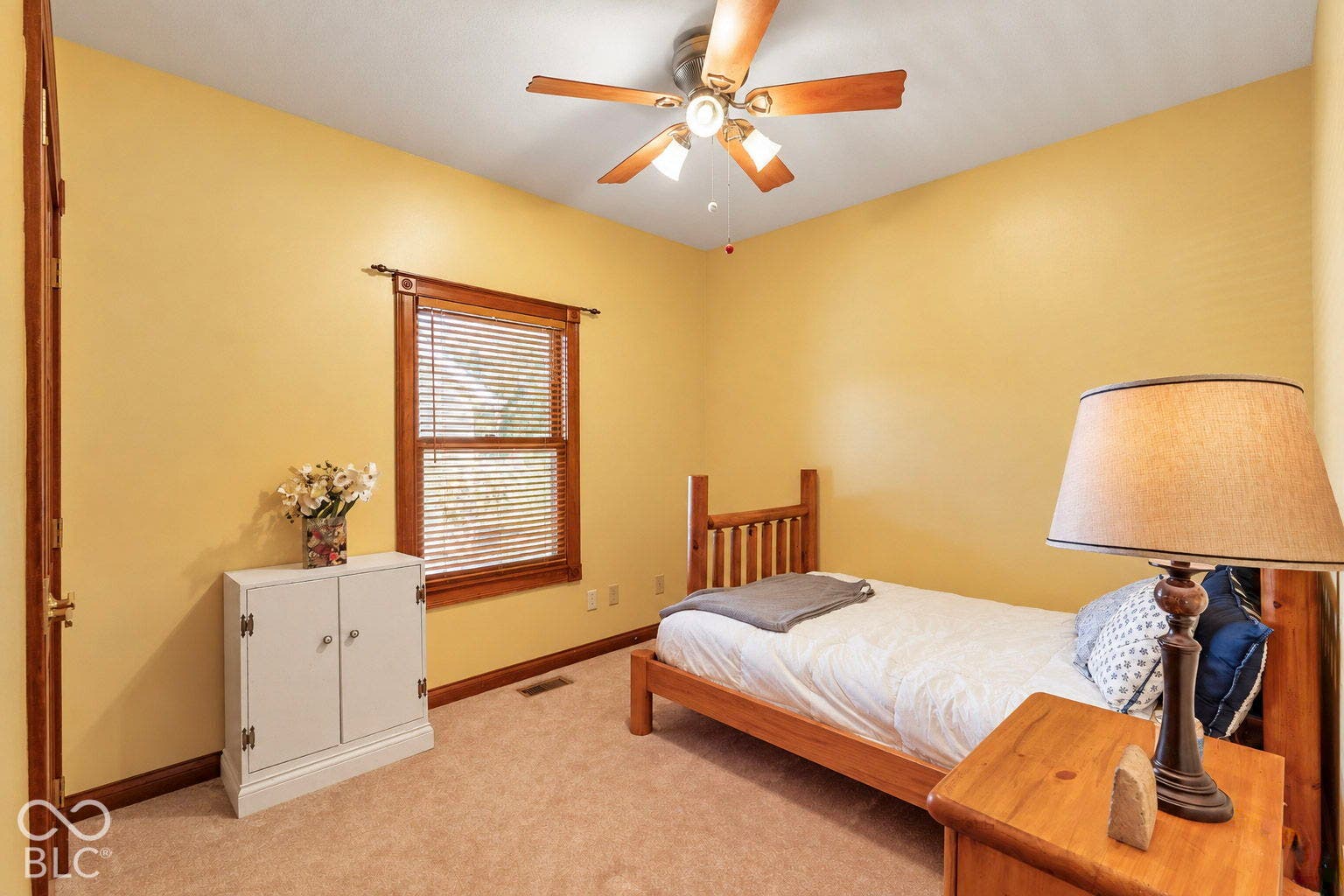 Light-colored carpeting, warm yellow walls, a window with wood trim, and a ceiling fan with integrated lighting