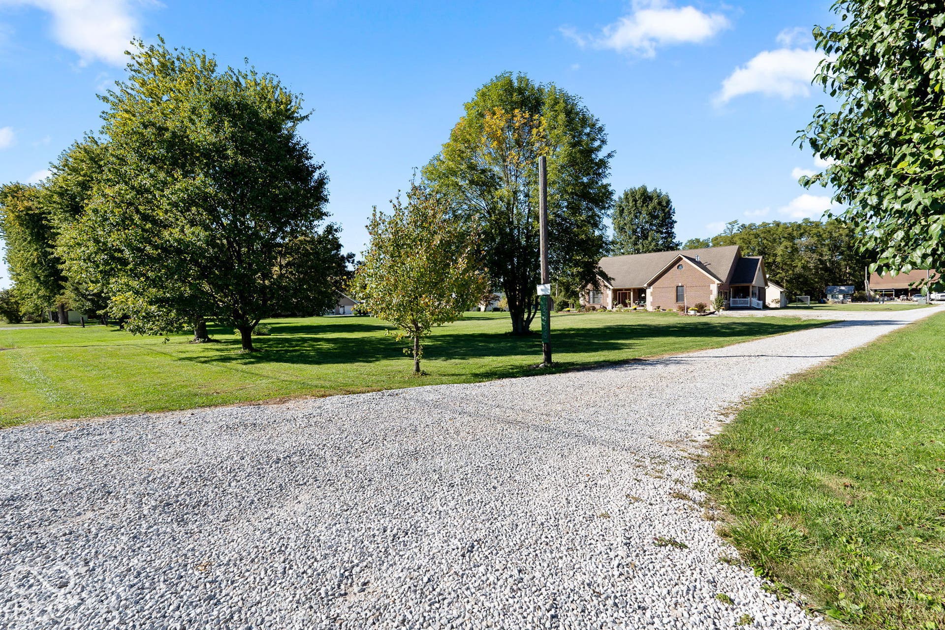 Gravel drive leading to a brick residence with a covered front porch, set on a manicured lawn with mature trees