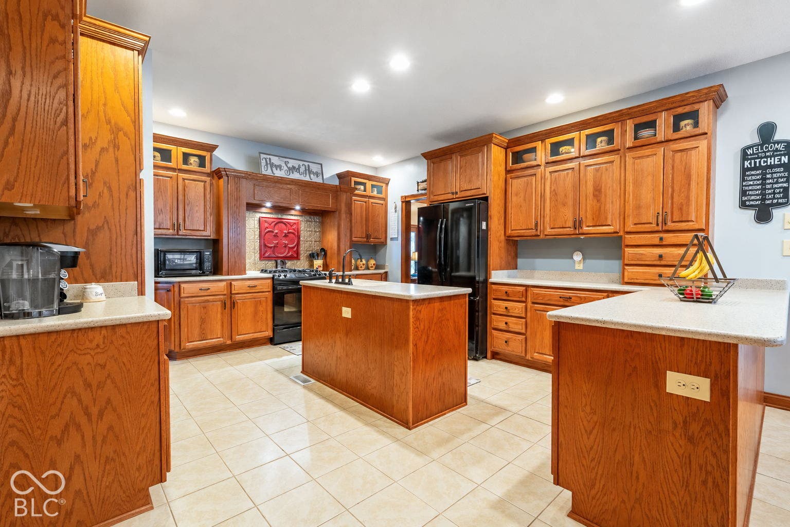 Kitchen featuring extensive wood cabinetry, light-toned countertops, a central island, tile flooring, and integrated black appliances