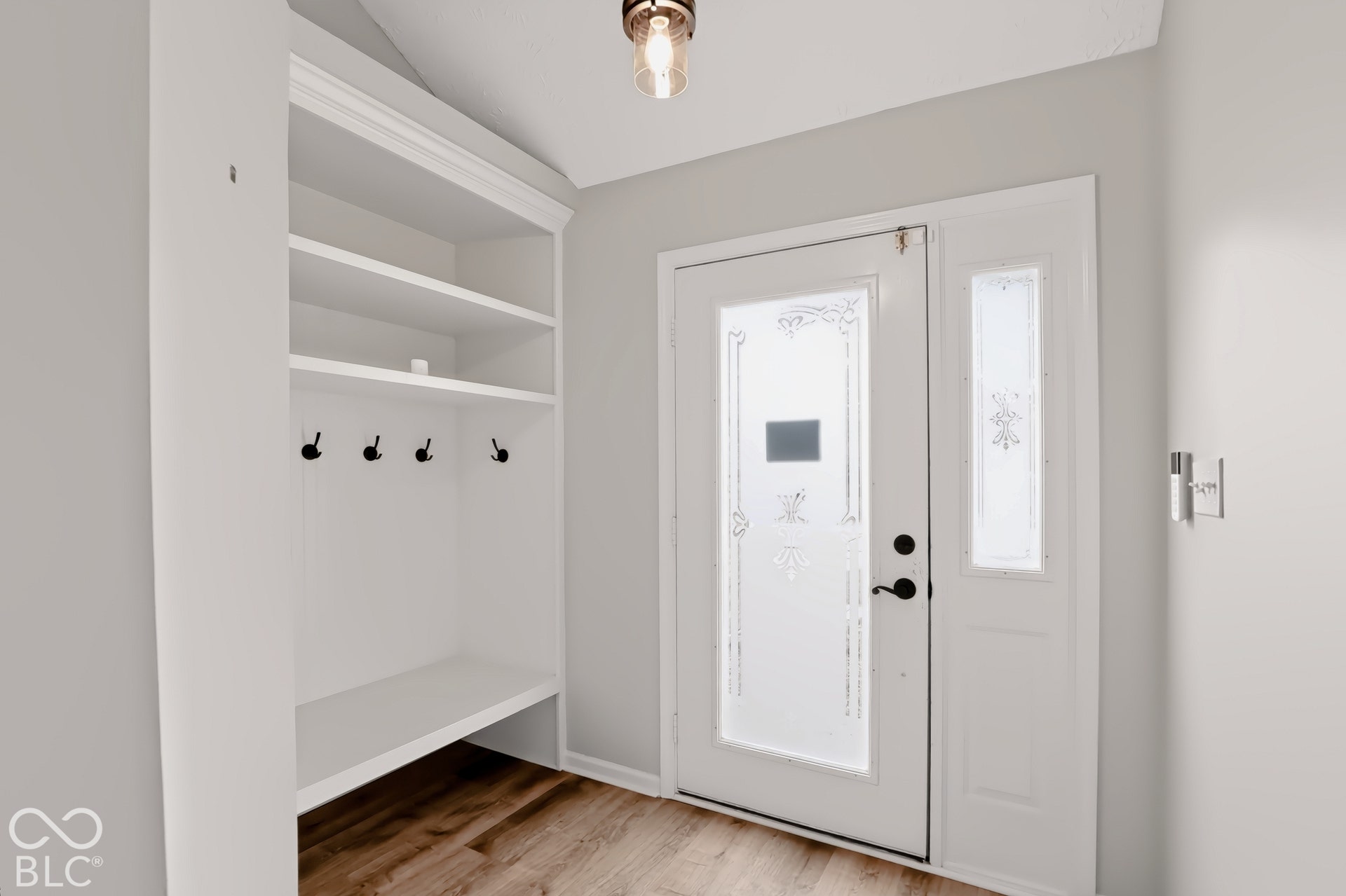 Bright entryway with white shaker-style cabinetry featuring open shelving and four integrated hooks, warm wood-finish flooring, recessed ceiling lighting, and direct access to the front entrance with sidelights and glass paneling