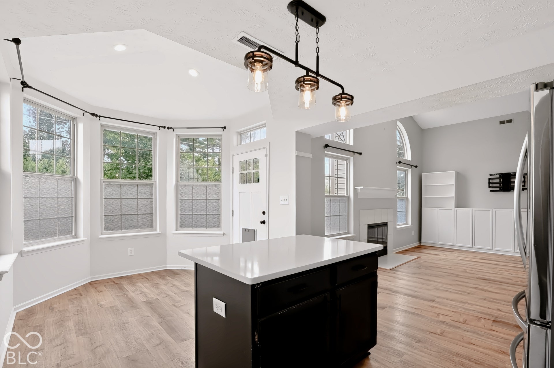 Open-concept living space featuring wood-finish flooring, a kitchen island with a light-toned countertop, a white paneled door with glass inserts, recessed lighting, and a fireplace with a white mantel and arched window