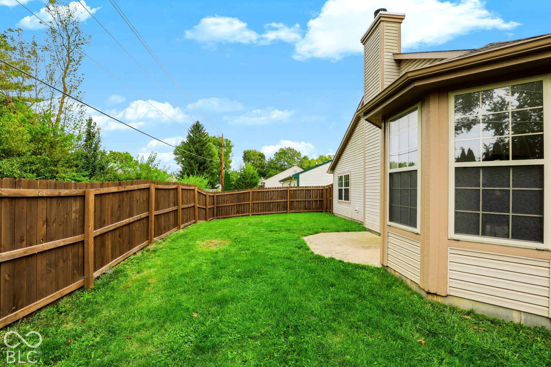 Expansive green lawn, privacy fencing, concrete patio, bay windows with grids, and a prominent exterior chimney
