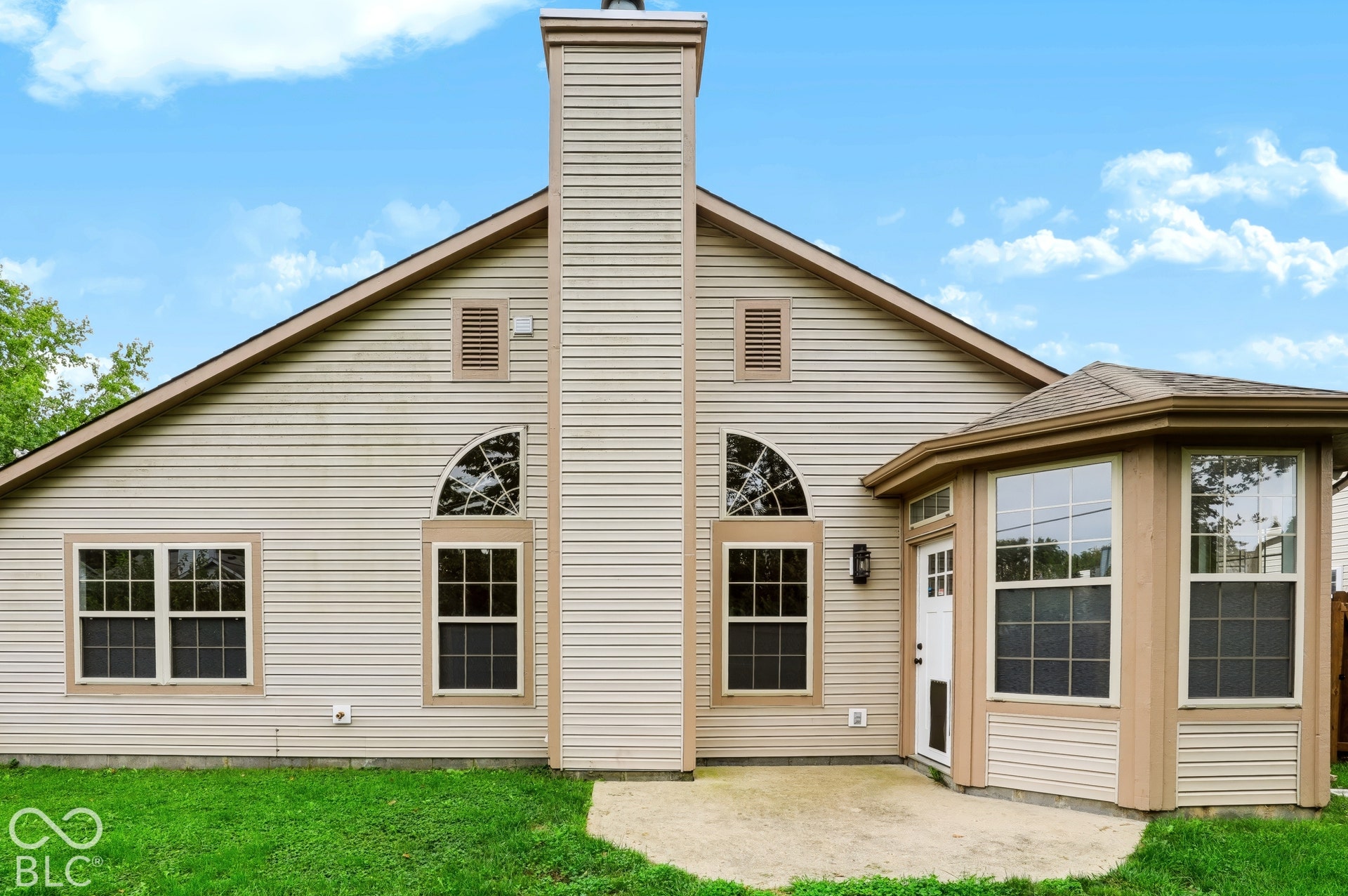 Exterior featuring beige siding, prominent chimney, bay window extension, arched transom windows, and a concrete patio