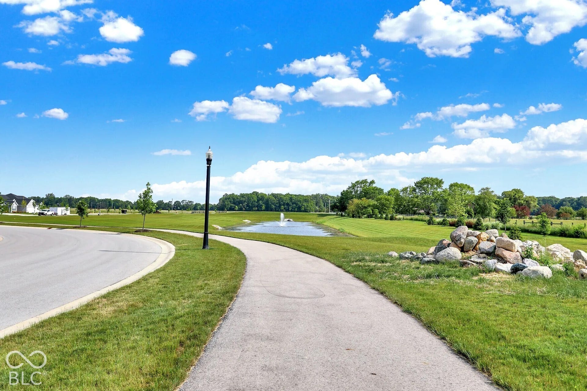 Paved walking path, street light fixture, rock landscaping feature, expansive green spaces, and a pond with a water feature