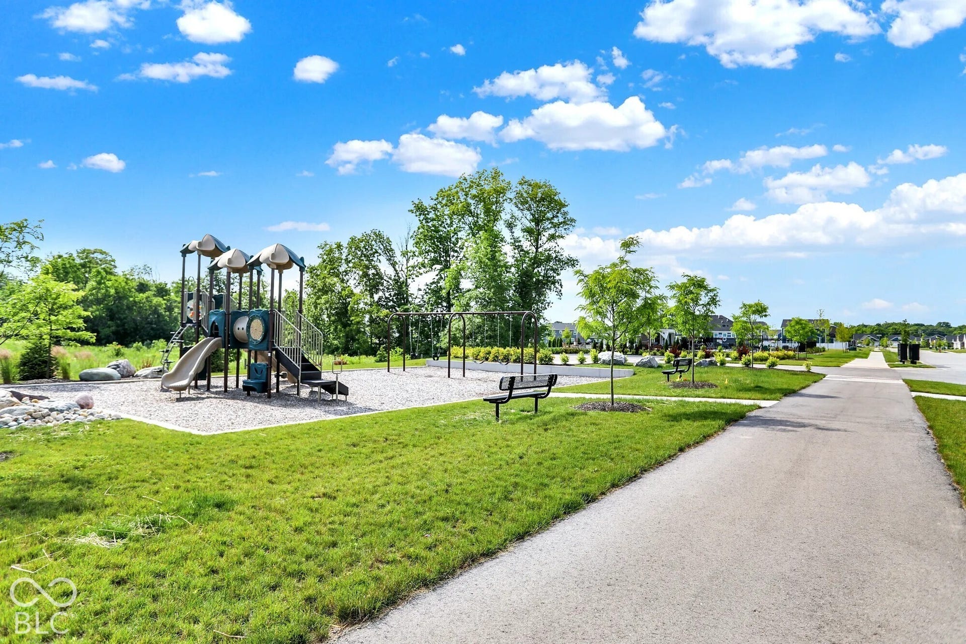 Playground with a multi-level structure and slides, adjacent to a paved walking path, surrounded by grass and trees
