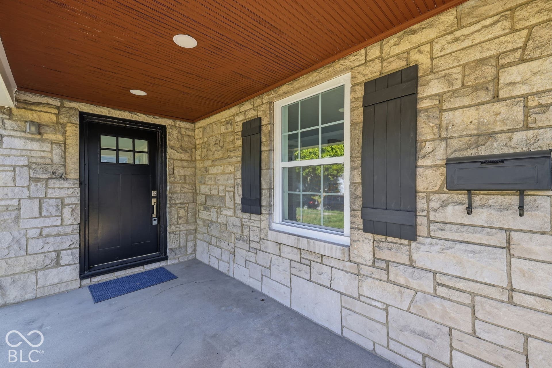 Stone veneer facade, dark paneled entry door with glass inserts, white window trim, dark exterior shutters, and a dark wall-mounted mailbox