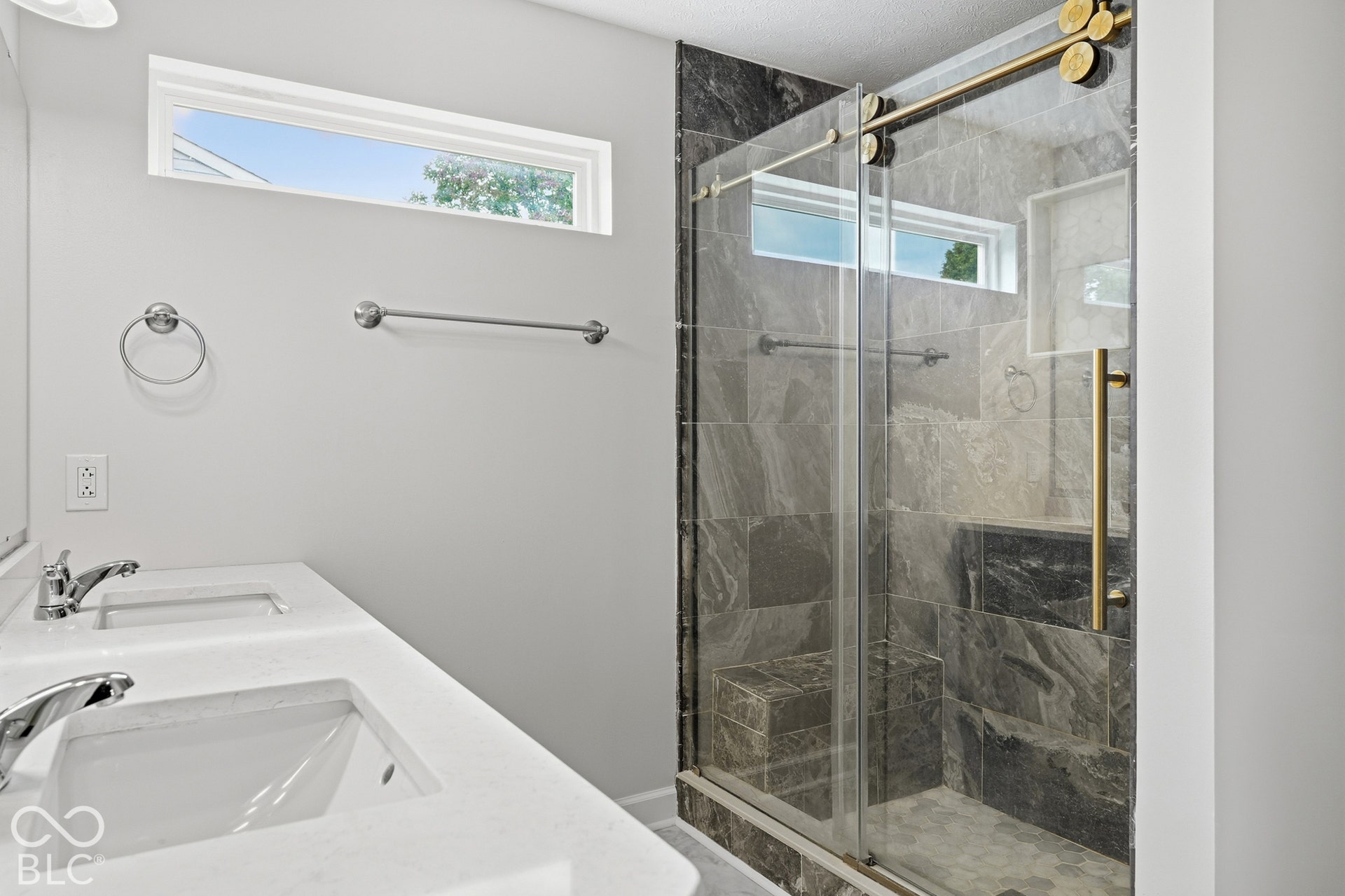 Dual vanity with integrated sinks and white countertops, complemented by a glass-enclosed shower featuring dark stone-look tile, a built-in shower bench, and gold-finish hardware