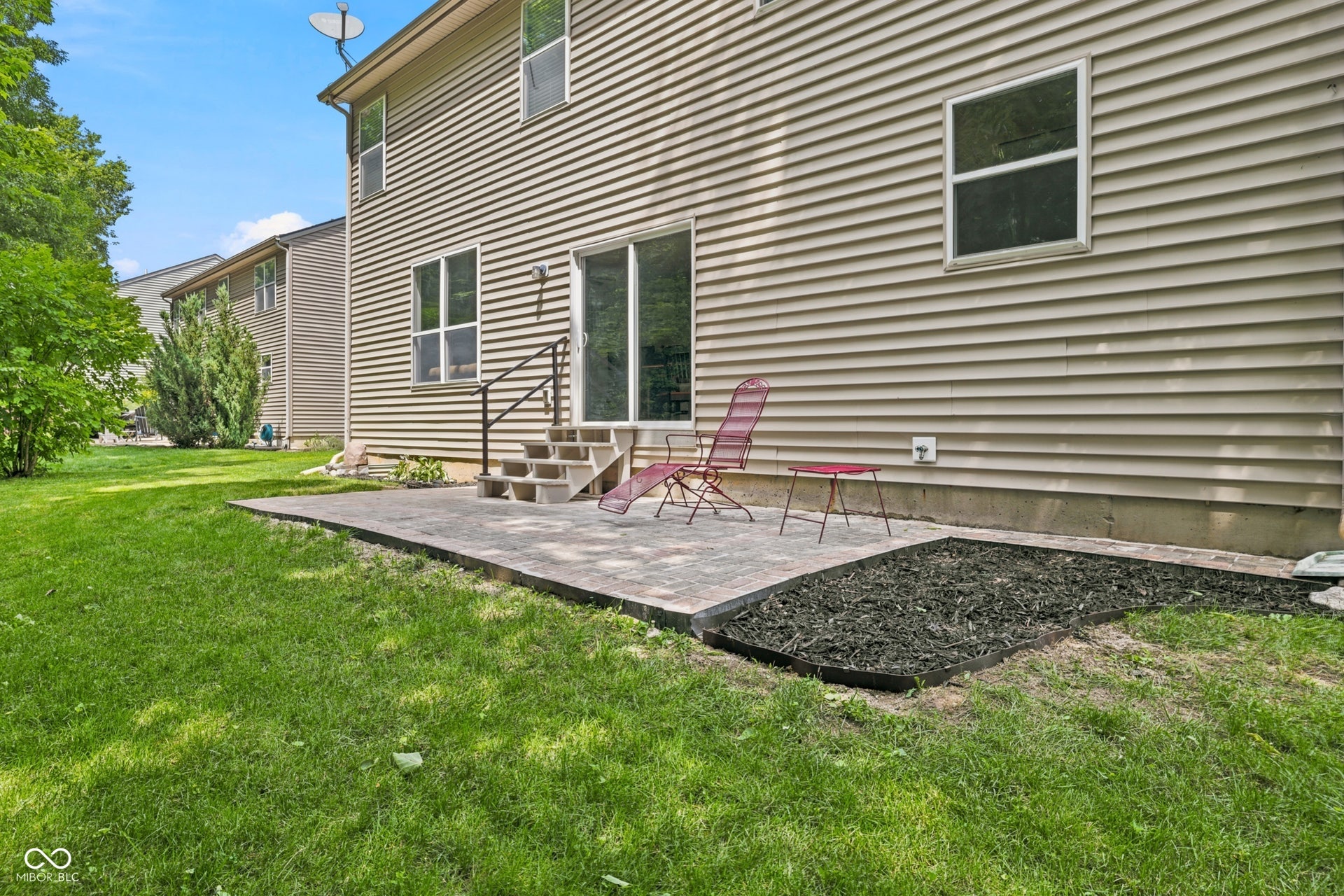 Rear exterior featuring a paver patio with steps, sliding glass door access, tan horizontal siding, and a mulched garden bed
