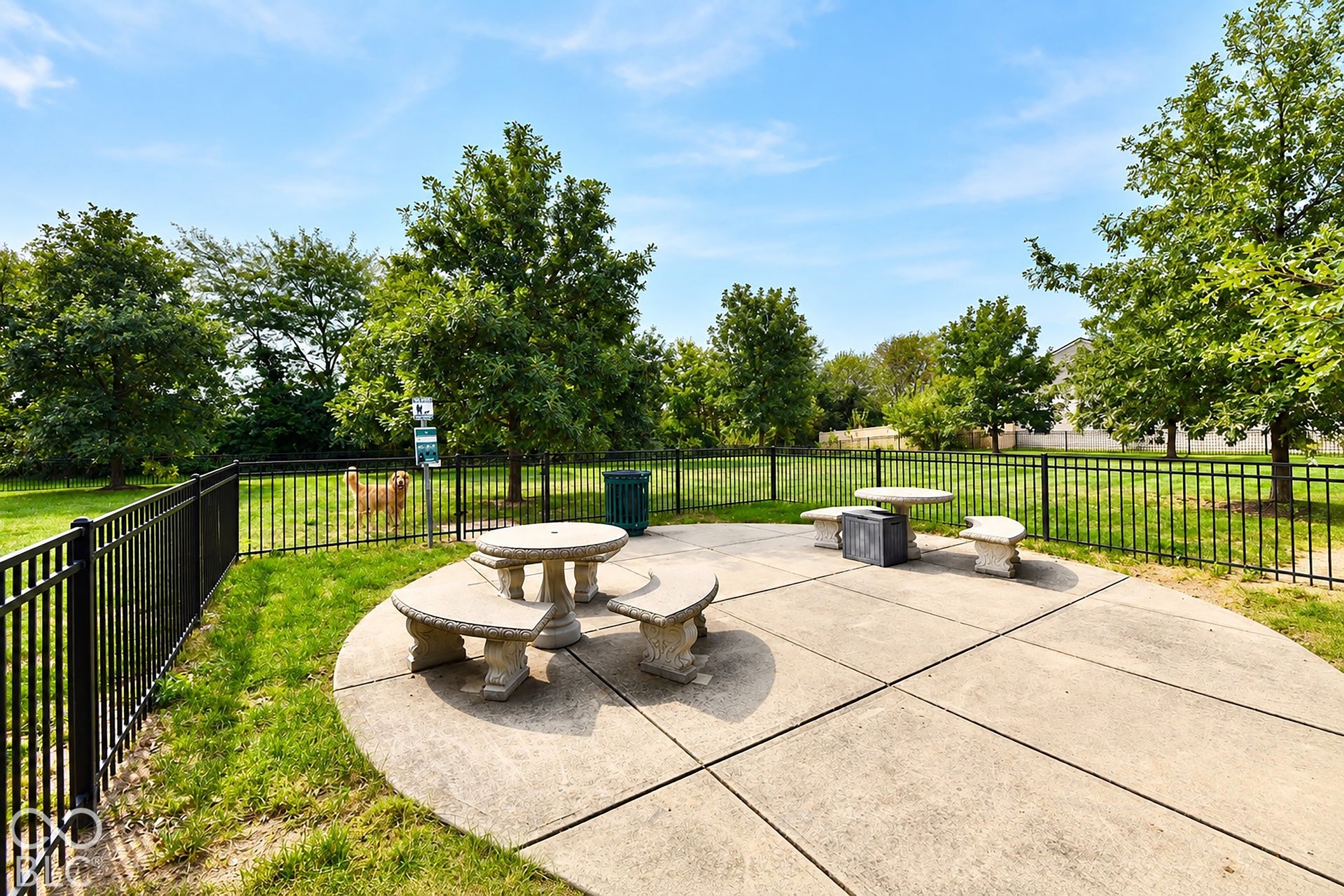 Neighborhood dog park with a Concrete patio with two permanent stone picnic tables, bordered by a black metal fence, and surrounded by mature trees