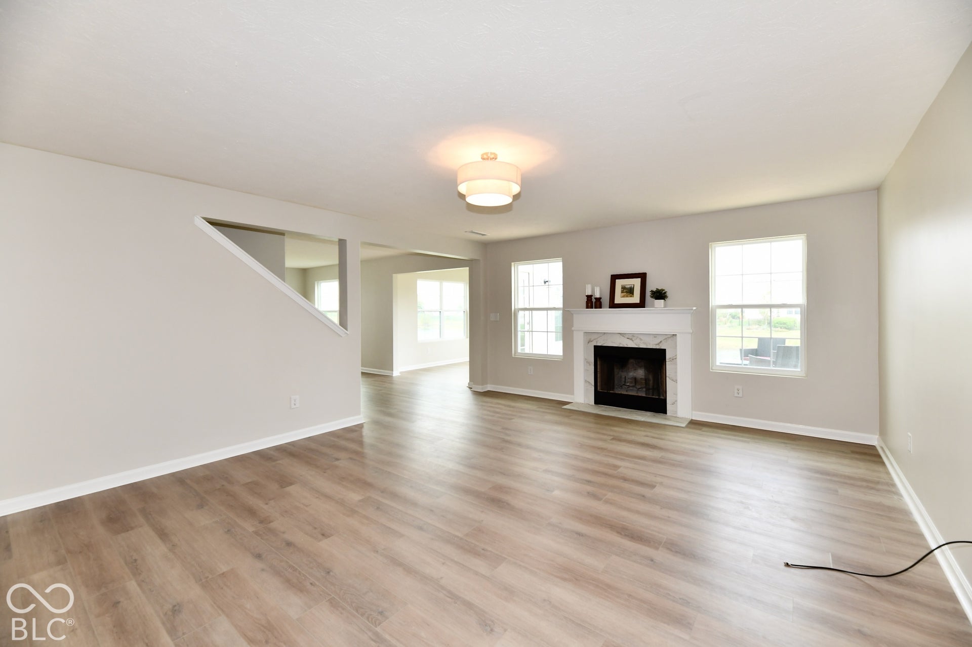 Expansive living area featuring light wood-finish flooring, a contemporary ceiling light fixture, a fireplace with a white mantel, and multiple windows