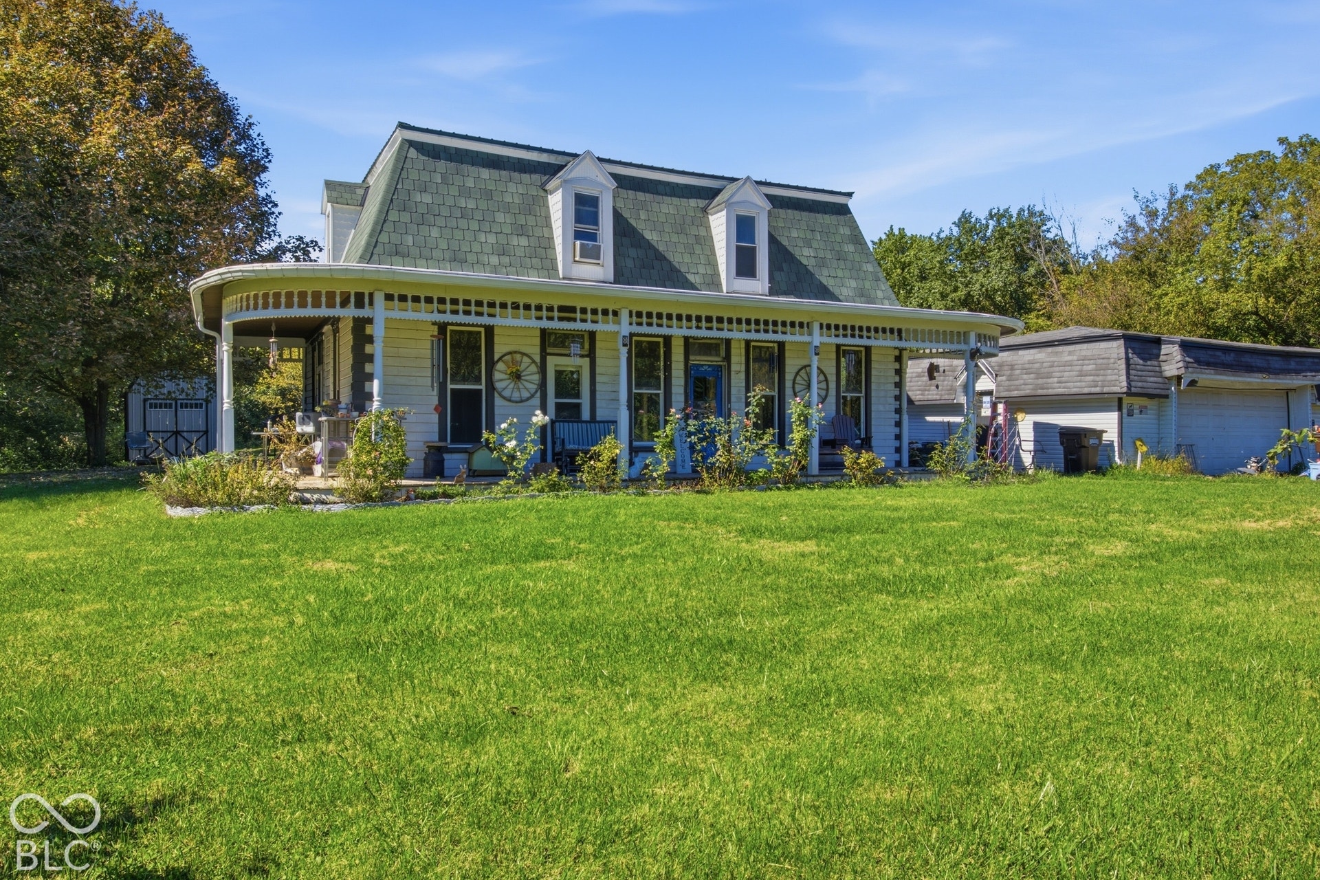 Expansive front lawn, wraparound porch with lattice detailing, gambrel roof, dormer windows, and an outbuilding