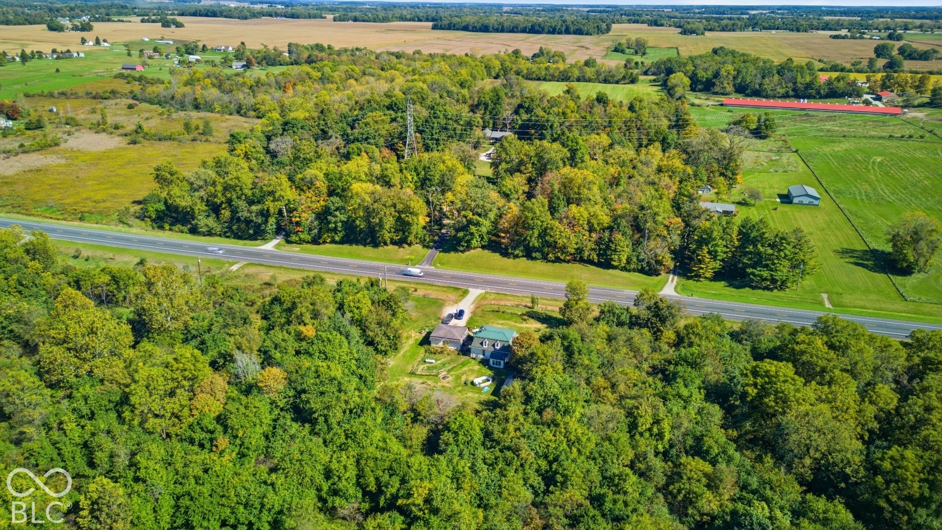 Expansive property featuring dense mature trees, a paved roadway, and open fields in the surroundings