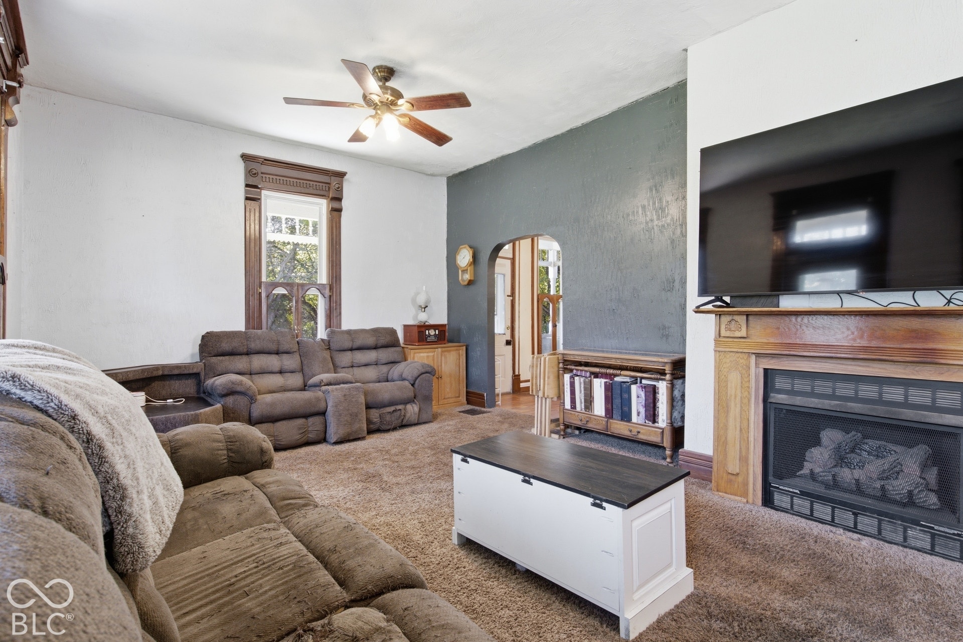 Spacious living room featuring a gas fireplace with wood surround, wall-mounted flat-screen television, ceiling fan, and plush seating arrangement