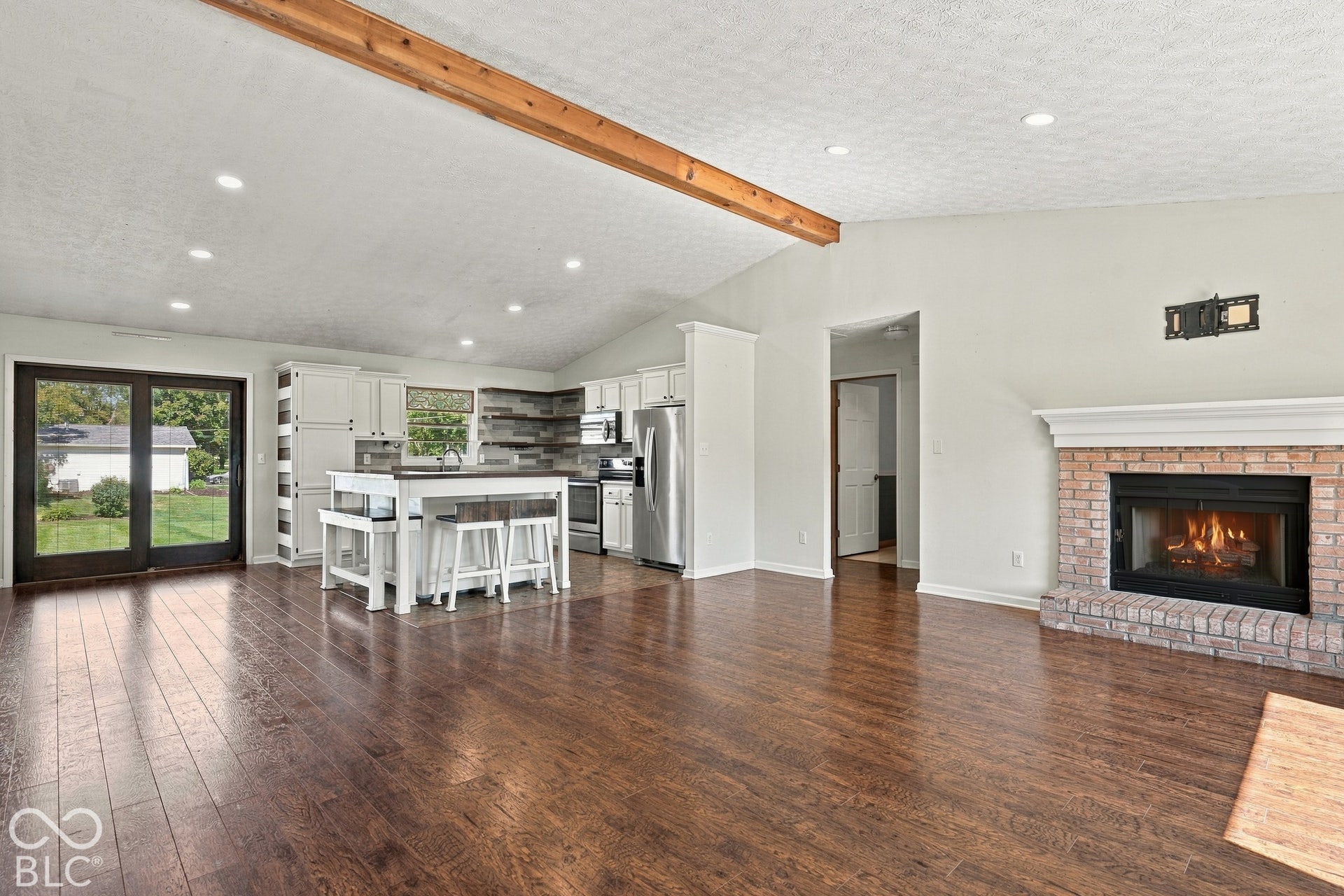 Vaulted ceiling with exposed wood beam, brick-surround fireplace with mantel, dark wood-finish flooring, white kitchen cabinetry with island, and glass patio doors