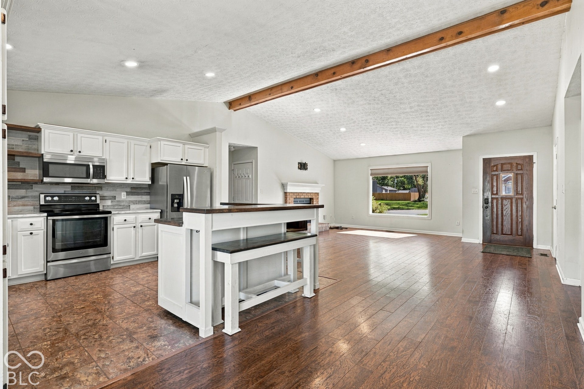 Open-concept living space featuring vaulted ceilings with exposed wood beams, wood-finish flooring, a kitchen island with seating, white shaker cabinetry, and stainless steel appliances