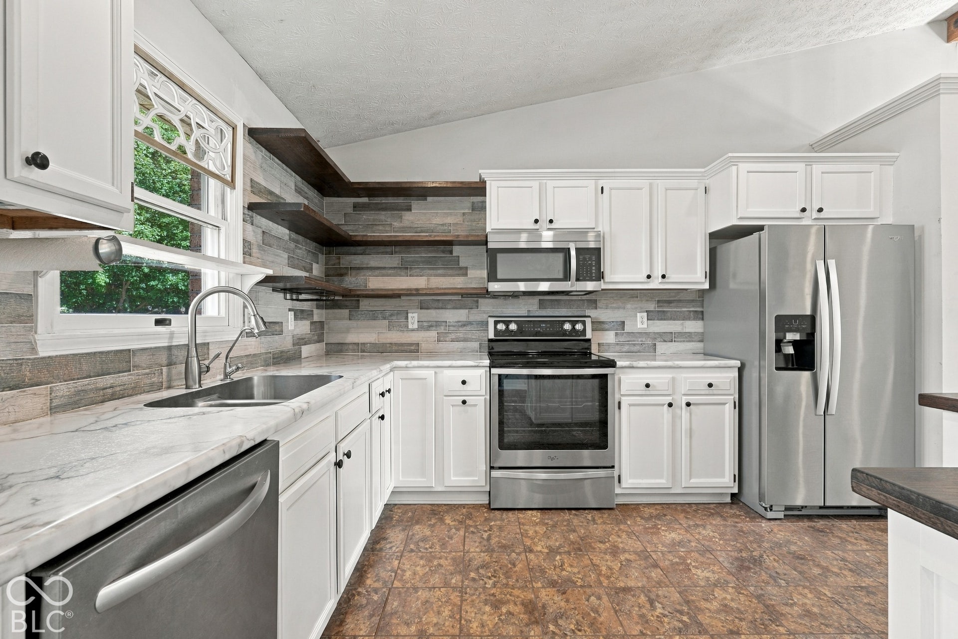 White cabinetry with dark hardware, stainless steel appliances, light-toned countertops, a textured backsplash, and open wood shelving