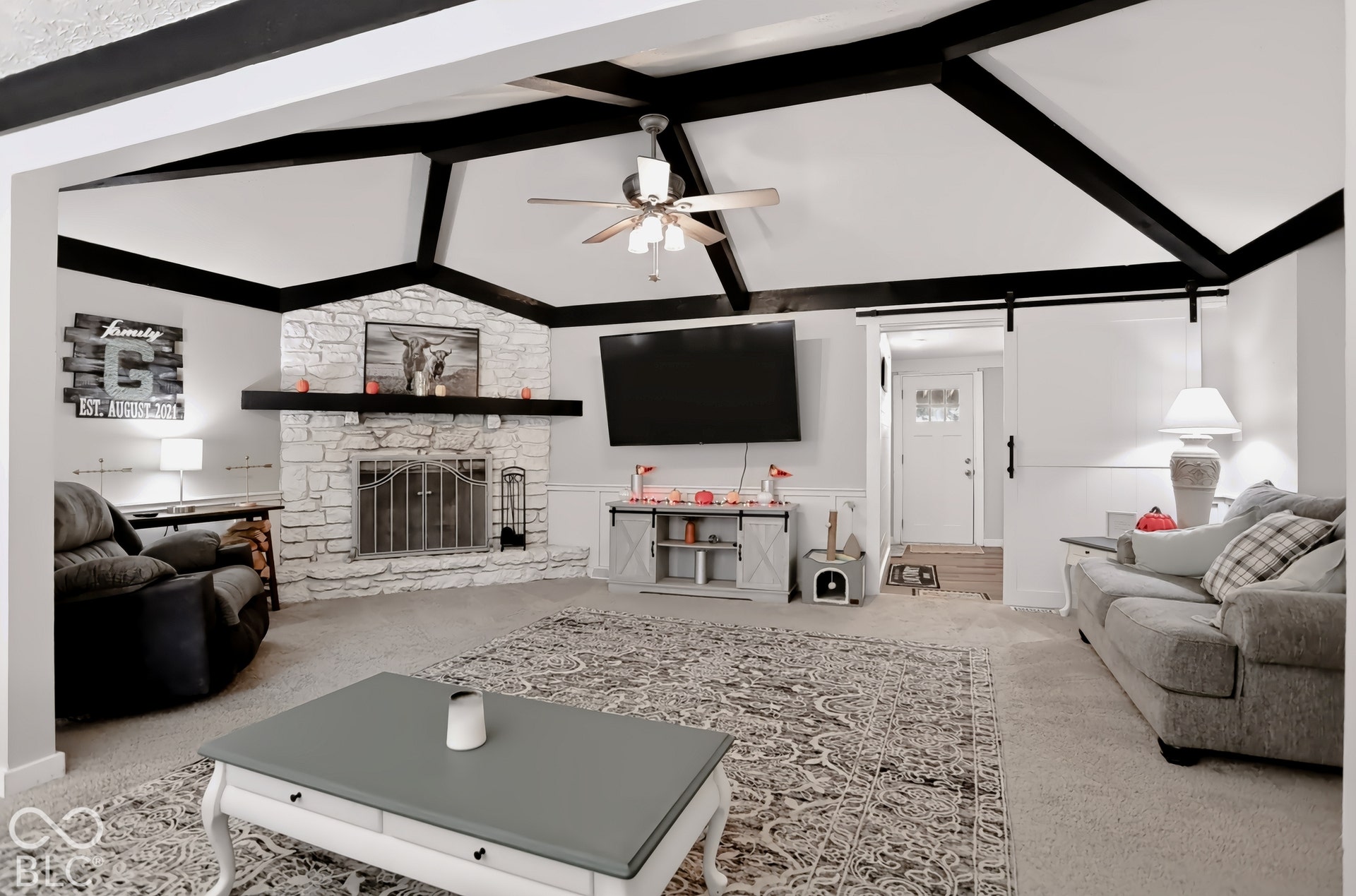 Vaulted ceiling with exposed beams, stone-clad fireplace with mantel, ceiling fan, and white wainscoting