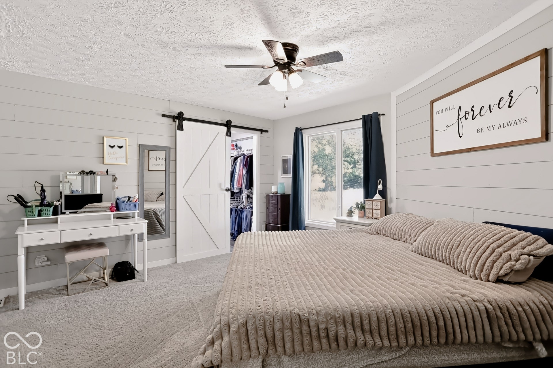 Shiplap accent wall, light-colored carpeting, ceiling fan with light fixture, large window with dark drapes, and white barn-style sliding closet doors