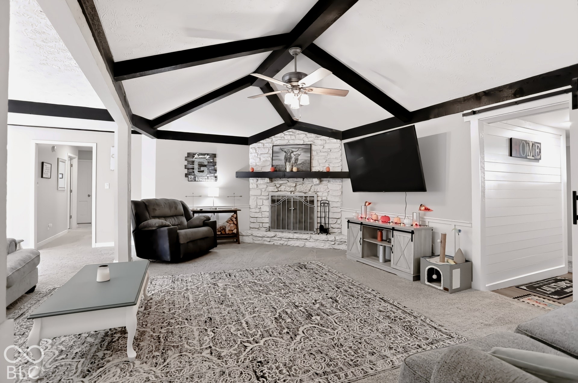 Vaulted ceiling with exposed beams, stone-clad fireplace with mantel, ceiling fan with light fixture, carpeted flooring, and a sliding barn door